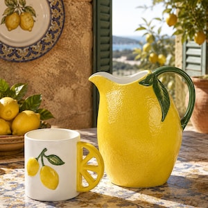 Può includere: Una brocca in ceramica a forma di grande limone giallo con manico verde e dettaglio a foglia, abbinata a una tazza bianca con un grafico di limone e un manico giallo a forma di spicchio di agrume, entrambe con superfici testurizzate.