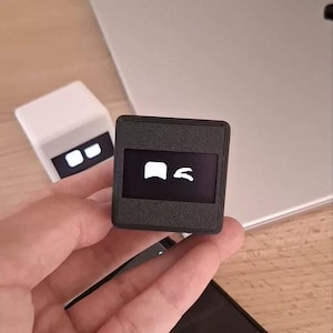 May include: A small, square, black electronic device with a textured finish and a front-facing digital screen displaying a white icon of a square and a curved shape. A similar white, smaller device is visible in the background.