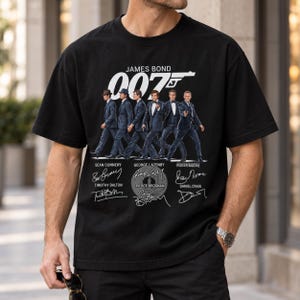 Puede incluir: Una camiseta negra de manga corta con un gráfico de seis actores de James Bond en traje, el logotipo 007 con una pistola, y los nombres y firmas impresos de Sean Connery, George Lazenby, Roger Moore, Timothy Dalton, Pierce Brosnan y Daniel Craig.