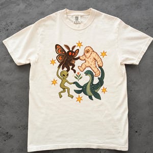 May include: An off-white short-sleeve t-shirt featuring a graphic print of a moth, a yeti, an alien, and a sea creature holding hands in a circle around a small flower, surrounded by yellow stars. The shirt has a Comfort Colors brand label.