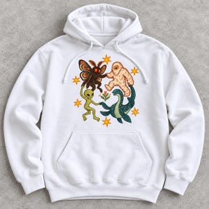 Op de afbeelding: Een witte hoodie met een centrale afbeelding van een mot, een yeti, een alien en een zeewezen die elkaars hand vasthouden in een cirkel rond een kleine bloem, omringd door verschillende oranje vierpuntige sterren.
