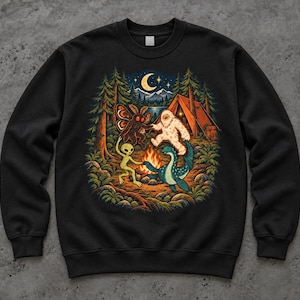 May include: A black crewneck sweatshirt featuring a graphic illustration of a mothman, a yeti, an alien, and a sea monster dancing around a campfire in a forest at night with a tent, mountains, and a crescent moon in the background.
