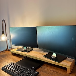 Op de afbeelding: Twee zwarte BenQ-computermonitoren staan op een lichtgekleurde houten monitorstandaard. Een zwarte ThinkPad-laptop is onder de standaard geplaatst, met een zwart Logitech-toetsenbord en een zwarte computermuis op het houten bureauoppervlak.