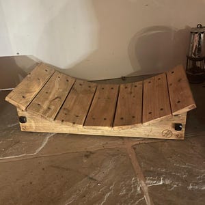 May include: A curved wooden log holder constructed from six horizontal planks secured with dark metal screws. The light brown wood features a visible grain and a circular brand stamp on the lower right side that reads Rustic Designs.