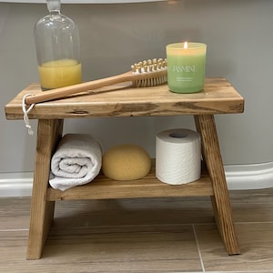 May include: A rustic wooden stool with two tiers. The top shelf holds a clear glass bottle with yellow liquid, a long-handled wooden bath brush, and a lit green candle labeled Jasmine. The bottom shelf holds a white towel, a yellow sponge, and toilet paper.