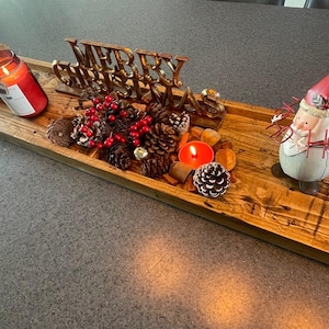 May include: A long rectangular wooden tray holding a gold Merry Christmas sign, a red candle in a glass jar, a small nativity scene, pinecones, red berries, a small orange tea light candle, and a white Santa figurine with red hat and Ho Ho Ho text.