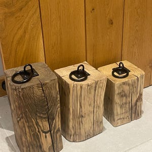 May include: Three rectangular wooden blocks of varying heights, each featuring a dark brown, weathered wood grain texture and a black metal ring handle securely mounted to the top surface with a square metal plate, creating a functional set.