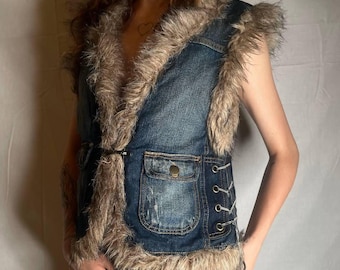May include: A sleeveless denim vest featuring a dark blue wash with distressed detailing. The vest is trimmed with long, shaggy faux fur along the collar, armholes, and hem. It includes two front pockets with metal buttons and side lace-up details.