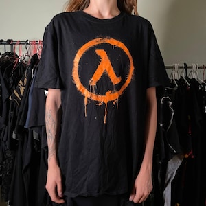 Puede incluir: Una camiseta negra de manga corta con un gráfico grande y desgastado de color naranja del símbolo Lambda en el pecho. El diseño del logotipo circular incluye efectos de goteo de pintura que se extienden hacia abajo desde el borde inferior del anillo.