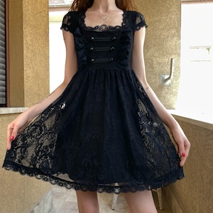 May include: A black knee-length dress featuring intricate lace detailing throughout the fabric. The garment has short cap sleeves, a square neckline, and a bodice accented with three horizontal black fabric tabs and matching buttons down the center.