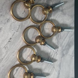 May include: A set of five brass-toned metal ring pulls, each featuring a circular ring attached to a central knob with a threaded silver-coloured metal bolt extending from the back for mounting purposes. The hardware has a polished finish.