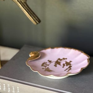 May include: A light pink ceramic dish shaped like a shell with a scalloped edge and a gold-painted rim. The center of the dish features a gold-colored botanical illustration of branches and leaves. The dish has a small, raised gold accent on one side.