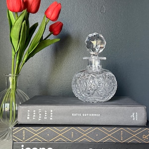 May include: A clear, round, cut-glass perfume bottle with a faceted spherical stopper, resting on a stack of three books titled More Than You'll Ever Know, A 1000 Better, and Icons by Oscar, alongside a glass vase containing four red tulips.