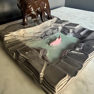 Pode incluir: Uma escultura de cerâmica com um urso castanho brilhante de pé sobre uma formação rochosa cinzenta e estratificada que rodeia um lago central. Um pequeno peixe cor-de-rosa está posicionado no centro da superfície de água azul-clara, lisa e brilhante.