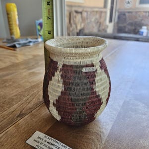 May include: A woven basket with a bulbous shape, featuring a geometric pattern in cream, dark brown, and deep red. A yellow measuring tape shows the basket stands approximately 3.5 inches tall. A small label reads 11/15 and another reads Woven basket and plates.