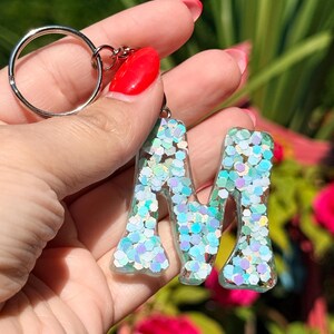 May include: A clear resin keychain in the shape of the letter "M", filled with iridescent glitter. The keychain has a silver metal ring attached. The background is blurred with green and pink hues.