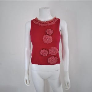 May include: A sleeveless red knit top featuring a ruffled neckline and four dimensional fabric flower appliqués in a lighter shade of pink. The flowers are arranged asymmetrically on the front of the garment, which is displayed on a mannequin.