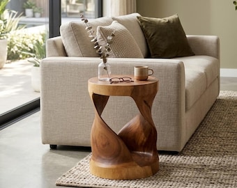 May include: A round wooden side table with a twisted, hourglass-shaped base featuring a smooth, polished finish. The table surface holds a clear glass vase with a dried plant stem, a pair of dark-rimmed eyeglasses, and a small, solid-coloured ceramic mug.