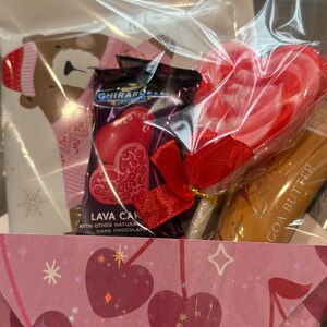 Puede incluir: Una cesta de regalo envuelta en celof&aacute;n transparente. Contiene una piruleta en forma de coraz&oacute;n, chocolates Ghirardelli Lava Cake y un tubo de manteca de cacao. La cesta tambi&eacute;n incluye una tarjeta con una ilustraci&oacute;n de oso y un sobre rosa con motivos de coraz&oacute;n.