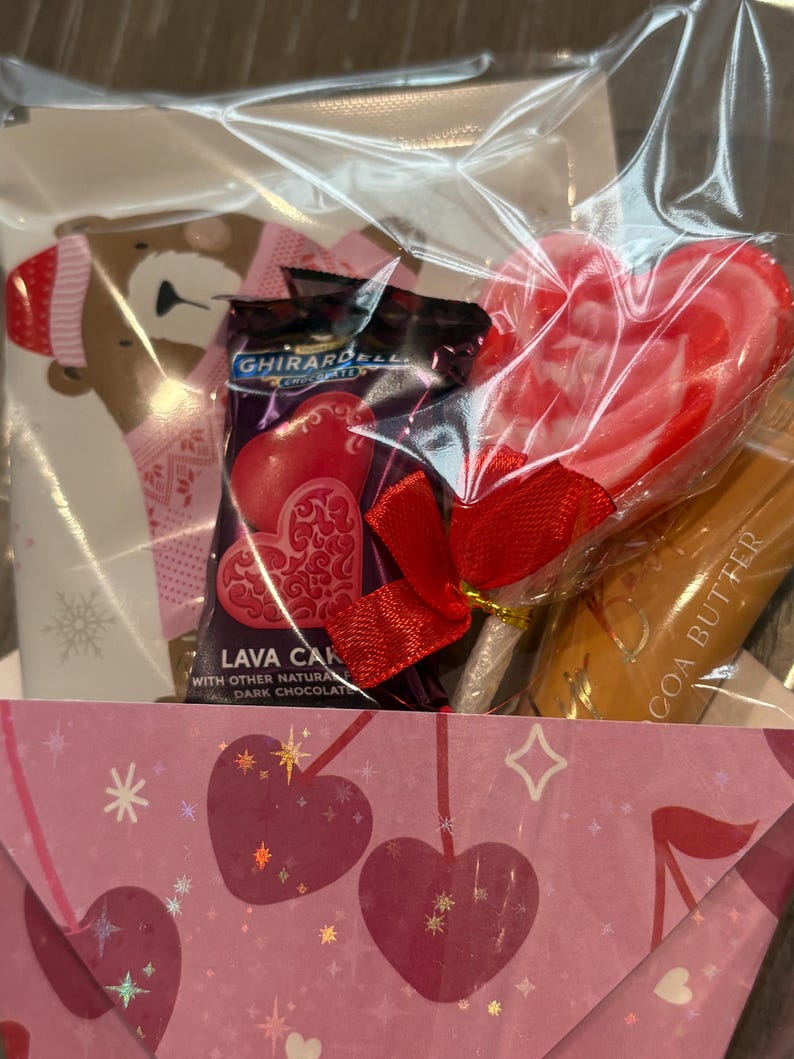Puede incluir: Una cesta de regalo envuelta en celof&aacute;n transparente. Contiene una piruleta en forma de coraz&oacute;n, chocolates Ghirardelli Lava Cake y un tubo de manteca de cacao. La cesta tambi&eacute;n incluye una tarjeta con una ilustraci&oacute;n de oso y un sobre rosa con motivos de coraz&oacute;n.