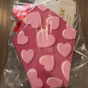 Puede incluir: Una bolsa de pl&aacute;stico transparente que contiene una mascarilla facial con motivos de corazones rosas y rojos y un peque&ntilde;o accesorio en forma de coraz&oacute;n rojo. La mascarilla tiene un patr&oacute;n de corazones rosas sobre un fondo rosa m&aacute;s oscuro. Una etiqueta con la palabra "SK" es visible.