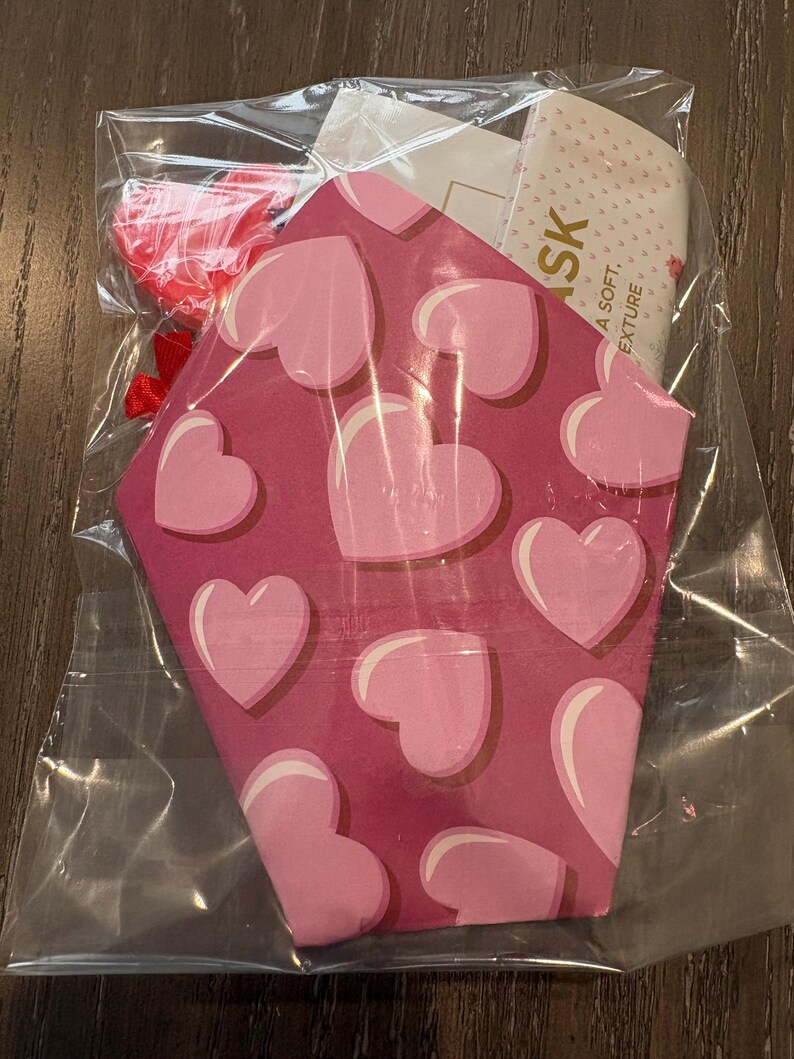 Puede incluir: Una bolsa de pl&aacute;stico transparente que contiene una mascarilla facial con motivos de corazones rosas y rojos y un peque&ntilde;o accesorio en forma de coraz&oacute;n rojo. La mascarilla tiene un patr&oacute;n de corazones rosas sobre un fondo rosa m&aacute;s oscuro. Una etiqueta con la palabra "SK" es visible.