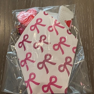 Puede incluir: Una bolsa de pl&aacute;stico transparente contiene una caja de papel blanca con un patr&oacute;n de lazos rosas. Un dije rojo en forma de coraz&oacute;n con una cadena dorada es visible. La caja tiene forma hexagonal.