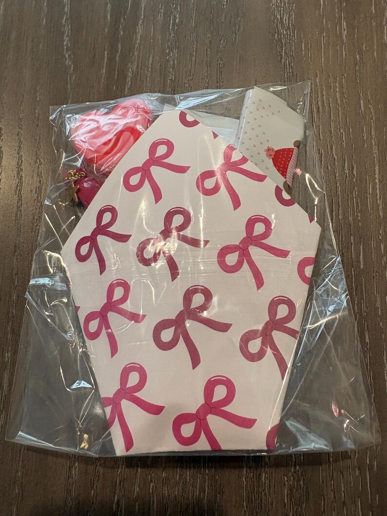 Puede incluir: Una bolsa de pl&aacute;stico transparente contiene una caja de papel blanca con un patr&oacute;n de lazos rosas. Un dije rojo en forma de coraz&oacute;n con una cadena dorada es visible. La caja tiene forma hexagonal.