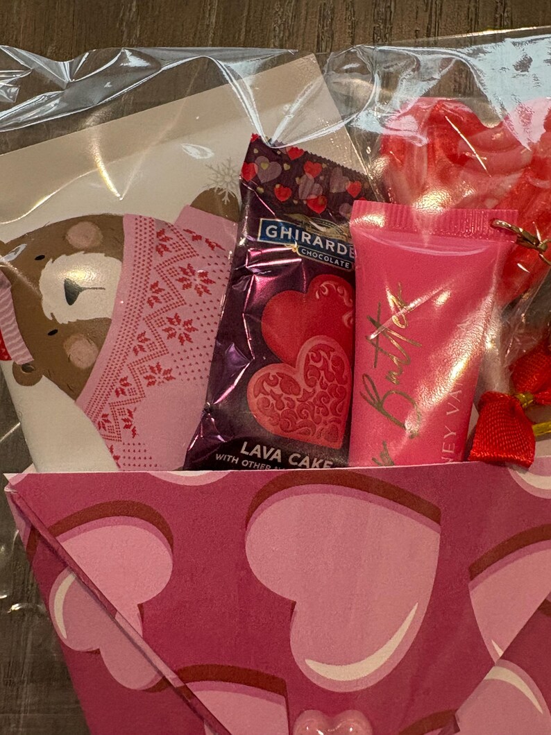 Puede incluir: Una cesta de regalo de San Valent&iacute;n envuelta en celof&aacute;n transparente. La cesta contiene una barra de chocolate Ghirardelli, caramelos en forma de coraz&oacute;n, un tubo de brillo labial rosa y una tarjeta decorativa.