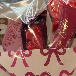 Puede incluir: Una cesta de regalo con tem&aacute;tica de San Valent&iacute;n. Contiene caramelos en forma de coraz&oacute;n, un tubo de manteca labial de fresa y una mascarilla facial con estampado rosa. La cesta est&aacute; decorada con lazos rosas.