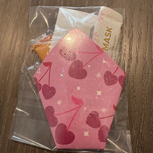 Puede incluir: Un paquete rosa en forma de coraz&oacute;n con tem&aacute;tica de cerezas en pl&aacute;stico transparente. El paquete contiene una mascarilla facial y un peque&ntilde;o accesorio naranja. El paquete tiene un patr&oacute;n de cerezas con la palabra "MASK" impresa.