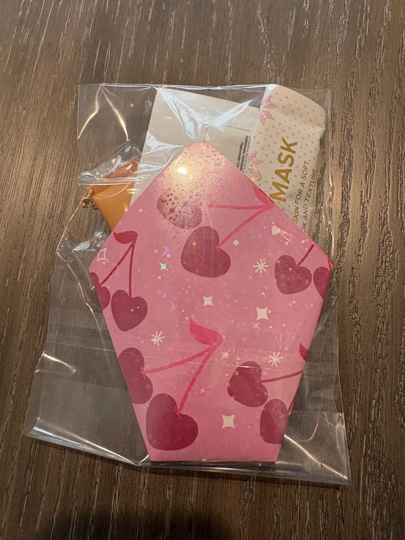 Puede incluir: Un paquete rosa en forma de coraz&oacute;n con tem&aacute;tica de cerezas en pl&aacute;stico transparente. El paquete contiene una mascarilla facial y un peque&ntilde;o accesorio naranja. El paquete tiene un patr&oacute;n de cerezas con la palabra "MASK" impresa.