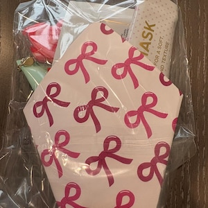 Puede incluir: Una bolsa de pl&aacute;stico transparente contiene un juego de regalo. El juego incluye una tarjeta estampada rosa y blanca con lazos rosas, un art&iacute;culo rosa, un art&iacute;culo verde y una mascarilla facial. La mascarilla facial tiene el texto "ASK FOR A SOFT, AND TEXTURE".