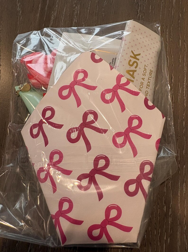 Puede incluir: Una bolsa de pl&aacute;stico transparente contiene un juego de regalo. El juego incluye una tarjeta estampada rosa y blanca con lazos rosas, un art&iacute;culo rosa, un art&iacute;culo verde y una mascarilla facial. La mascarilla facial tiene el texto "ASK FOR A SOFT, AND TEXTURE".