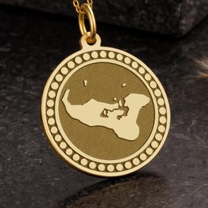 May include: A gold-toned circular pendant necklace featuring a map silhouette engraved on a matte olive-green centre. The pendant is framed by a decorative border of small raised dots and hangs from a delicate gold-toned chain link necklace.