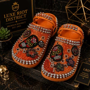 May include: A pair of orange clogs embellished with orange crystals and silver-toned metal spikes. Each clog features a sugar skull patch, a flower patch, and a circular emblem. The heel straps are also adorned with silver spikes and orange crystals.