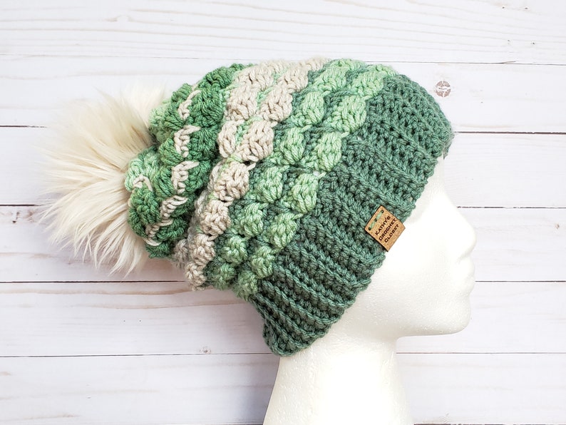 May include: A green, white and cream crocheted beanie with a fluffy white pom pom. The beanie has a brown leather tag that says "Kaitlynn's Crochet Glory".