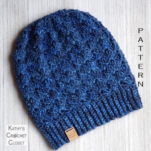 May include: Blue crocheted beanie hat with a textured stitch pattern. The hat has a fold-over brim and a brown leather tag with the text "Kathy's Crochet Closet".