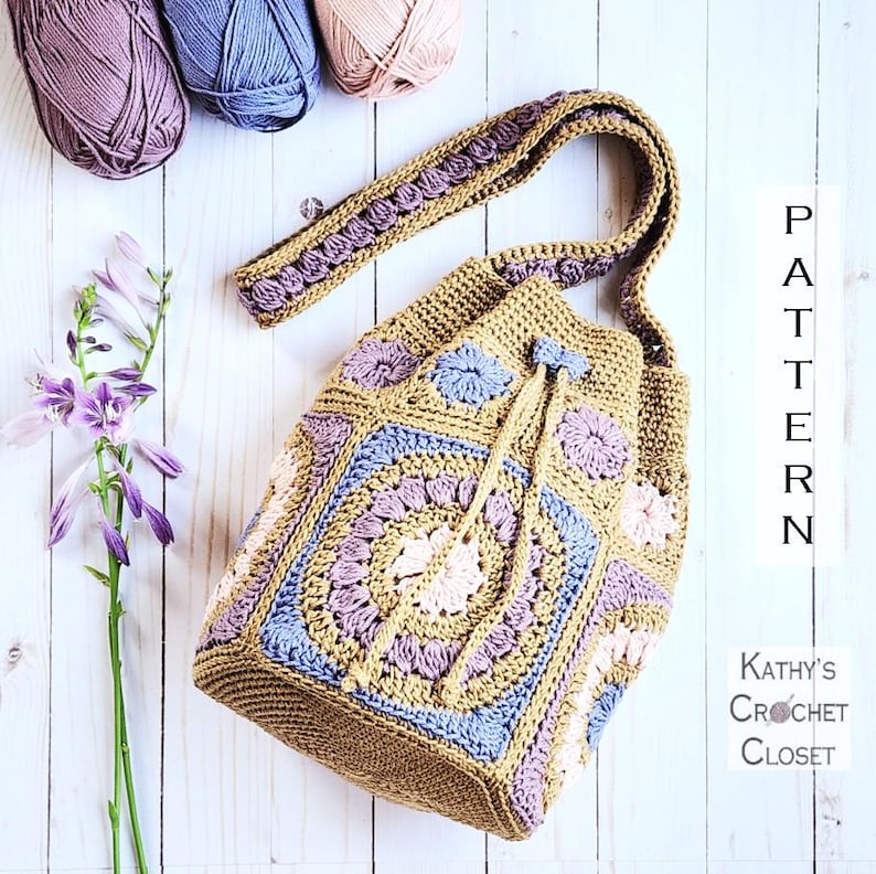 K&ouml;nnte beinhalten: Eine handgefertigte H&auml;keltasche in Beige-, Blau- und Lavendelt&ouml;nen. Die Tasche hat einen Kordelzugverschluss und einen Trageriemen. Das Design umfasst Blumen- und geometrische Muster. Das Bild zeigt auch Wollkn&auml;uel und den Text "PATTERN" und "Kathy's Crochet Closet".
