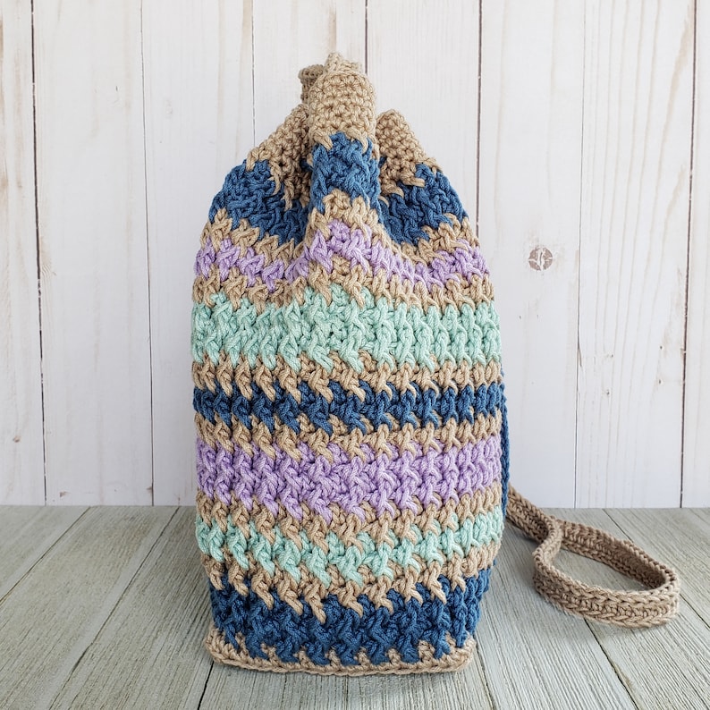 Pu&ograve; includere: Una borsa a cordoncino all'uncinetto con un motivo a righe in tonalit&agrave; di blu, verde, viola e marrone. La borsa ha una lunga tracolla in crochet marrone.