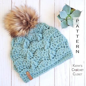 May include: A teal blue crocheted beanie with a brown faux fur pom pom. The beanie has a brown leather tag with the letters "KCC" on it. The beanie is on a white wooden surface. The text "PATTERN" is written vertically in white on a brown background to the right of the beanie. The text "Kathy's Crochet Closet" is written in black on a white background below the beanie.