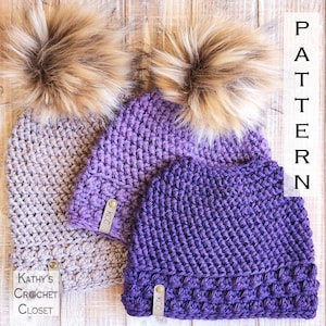 May include: Two crocheted hats with faux fur pom poms. One hat is gray and the other is purple with a gradient. Both hats have a leather tag with the letters "KCC" on them. The hats are on a wooden surface. The text "PATTERN" is written vertically on the right side of the image.