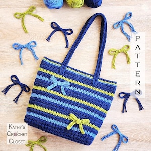 May include: A handmade crochet tote bag with blue, light blue, and yellow stripes. The bag features two blue handles and is decorated with small crochet bows in blue and yellow. The words "PATTERN" and "Kathy's Crochet Closet" are visible.