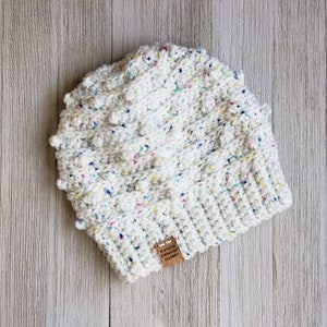 May include: White crochet beanie with colorful flecks and a textured bobble stitch. The beanie has a brown leather tag with the text "Katie's Crochet Closet".