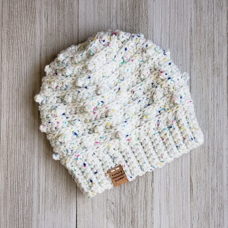 May include: White crochet beanie with colorful flecks and a textured bobble stitch. The beanie has a brown leather tag with the text "Katie's Crochet Closet".