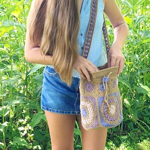 K&ouml;nnte beinhalten: Handgefertigte H&auml;keltasche mit Schulterriemen in den Farbt&ouml;nen Beige, Lila und Blau. Die Tasche hat einen Kordelzugverschluss und einen dekorativen Riemen. Das Design der Tasche umfasst Blumen- und geometrische Muster.