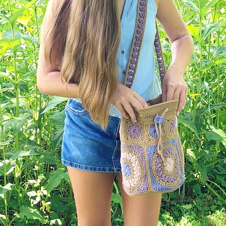 K&ouml;nnte beinhalten: Handgefertigte H&auml;keltasche mit Schulterriemen in den Farbt&ouml;nen Beige, Lila und Blau. Die Tasche hat einen Kordelzugverschluss und einen dekorativen Riemen. Das Design der Tasche umfasst Blumen- und geometrische Muster.