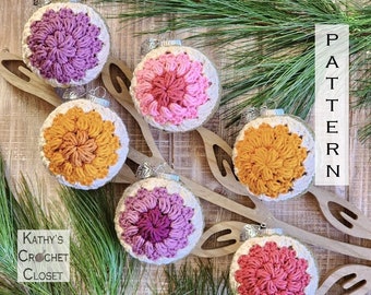 May include: Six crocheted ornaments in various shades of pink, purple, and orange. The ornaments are round and have a white background with a crocheted flower design in the center. The ornaments are hanging from a wooden branch with the text "PATTERN" written vertically on the right side of the image. The text "Kathy's Crochet Closet" is written in the lower left corner of the image.