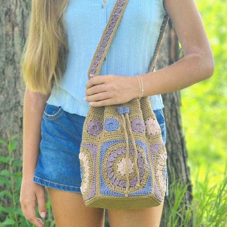 K&ouml;nnte beinhalten: Handgefertigte H&auml;keltasche mit Schulterriemen in Beige-, Lavendel- und Blaut&ouml;nen. Die Tasche hat einen Kordelzugverschluss und einen Riemen mit passendem H&auml;keldesign. Die Tasche ist mit floralen Motiven verziert.
