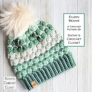 May include: A green, white and light green crochet beanie with a fluffy white pom pom. The beanie is made with a textured stitch and has a label that says "Kathy's Crochet Closet".